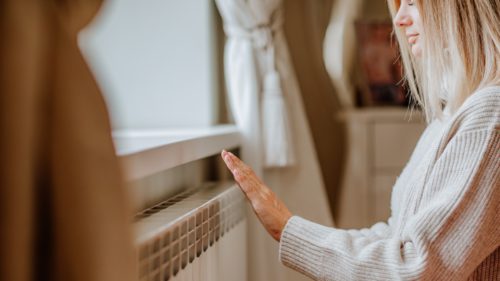 Young blond woman in long winter beige sweater is at home near the radiator. Winter season concept. Focus is at hands.