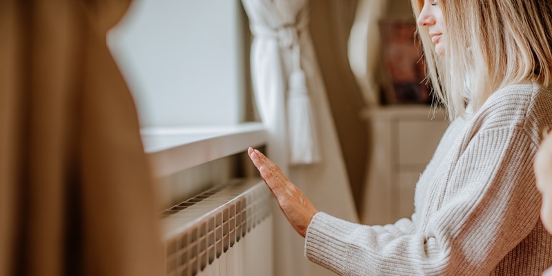 Young blond woman in long winter beige sweater is at home near the radiator. Winter season concept. Focus is at hands.