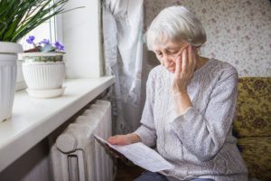 Worried, elderly woman examines her heating bill while sitting close to the radiator in winter