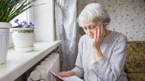 Worried, elderly woman examines her heating bill while sitting close to the radiator in winter