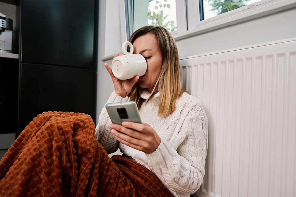 Young woman looks at her heating bill on her phone while sipping coffee and warming up by the radiator in winter